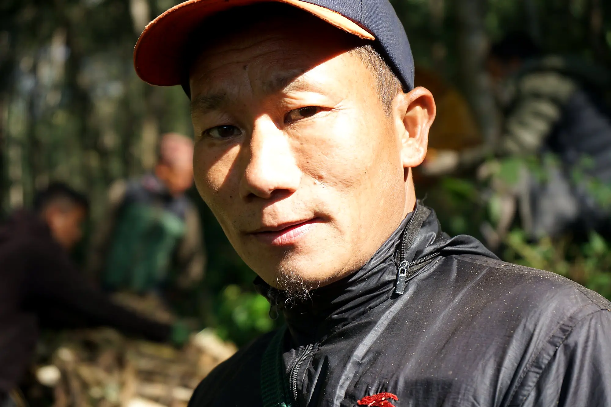 A close-up portrait of a young Nepali honey hunter wearing a navy and orange baseball cap and black zip-up jacket, looking directly into the camera with a composed expression, with a blurred forest background.