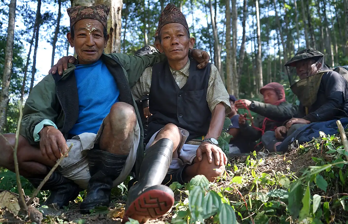 Two Gurung honey hunters wearing traditional embroidered Dhaka caps sit together in a sunlit forest smiling at the camera, with arms around each other, while other team members in protective bee veils rest in the background — representing the harvesting community behind Nepal's mad honey tradition.