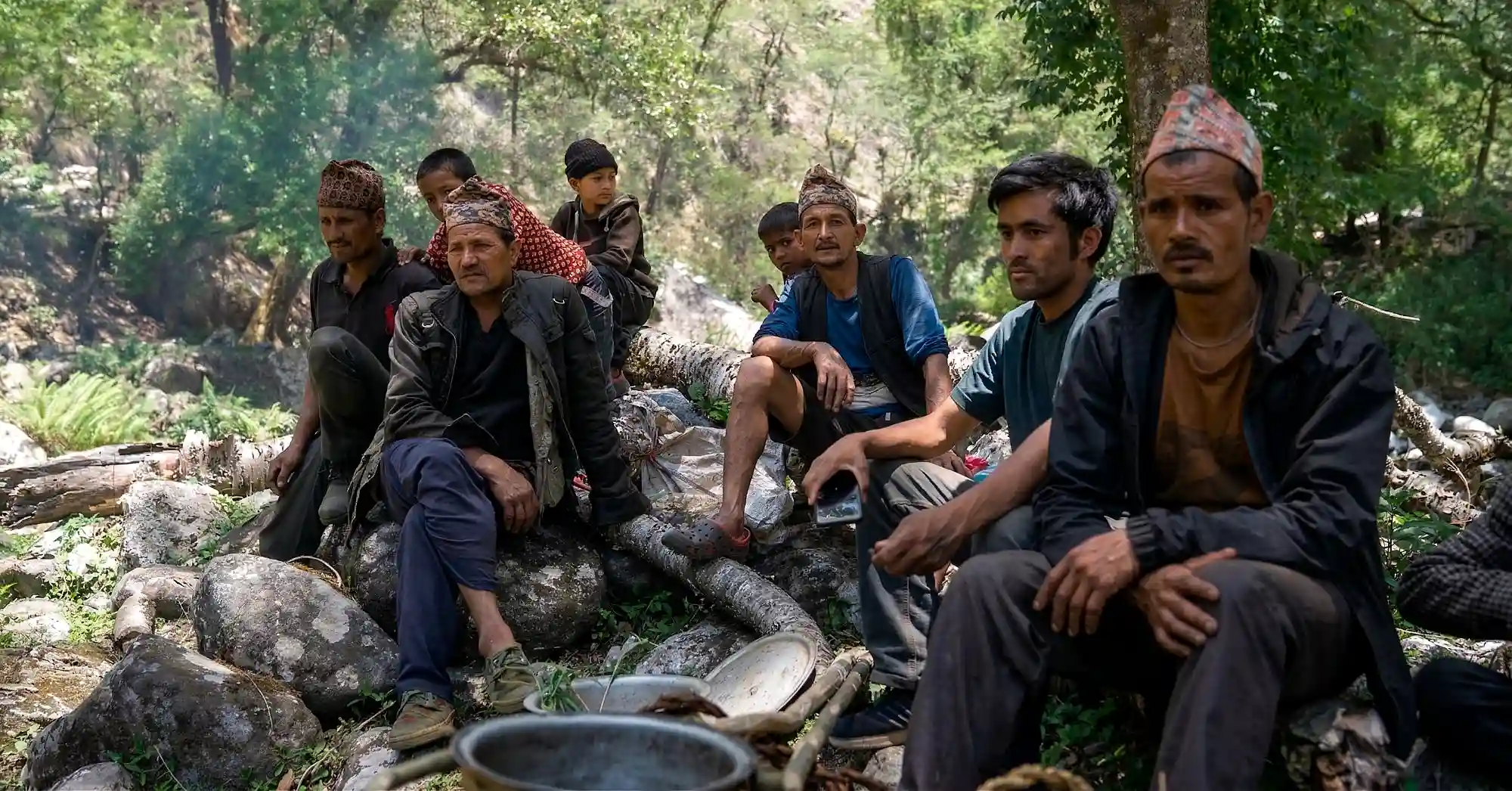 A group of Nepali Gurung honey hunters including men and boys wearing traditional Dhaka caps rest together on rocks and logs in a lush mountain forest beside harvesting equipment, between stages of a mad honey expedition.