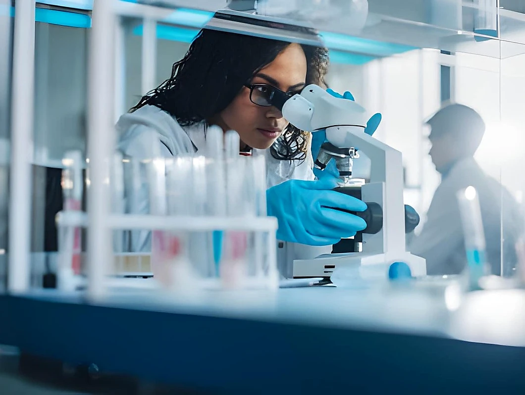A female scientist in a white lab coat and blue gloves examines a sample through a microscope in a modern laboratory, with rows of test tubes and lab equipment visible through glass panels in the foreground and a second researcher working in the background under blue laboratory lighting.