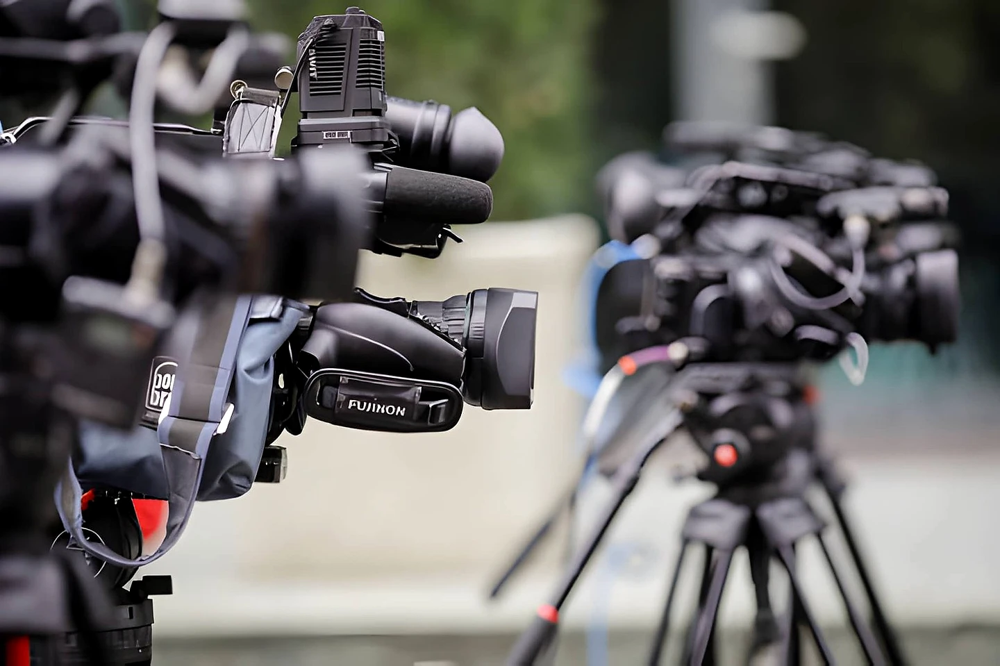 Multiple professional broadcast video cameras on tripods lined up at a media event, with Fujinon lenses in sharp focus in the foreground and additional cameras blurred in the background — representing press and documentary coverage.